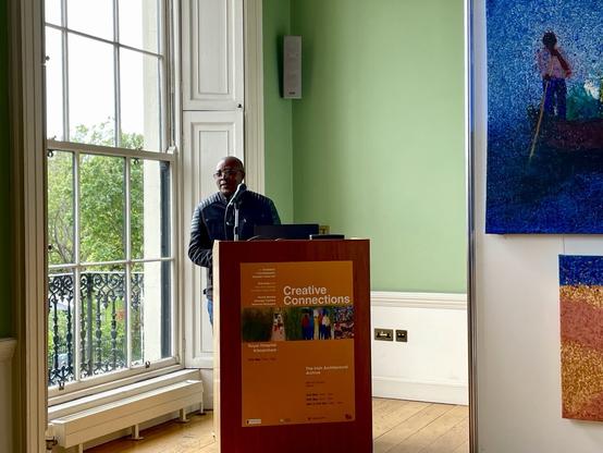 Patrick Mumba at lectern beside large sash window in heritage green walked room with wooden floorboards and his paintings partially in frame.