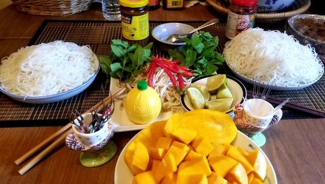 Rice noodles on two serving plates on table, on both sides with plate of bean sprouts, Vietnamese lemon mint, Thai hot chili peppers, lime wedges & small Realemon squeeze bottle. Crab forks are in two egg cups. A plate of sliced mango pieces are in front.
Prawn paste & garlic chili sauce containers are at back.