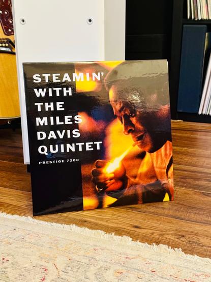 Photo of a record album jacket leaning against a white floorstanding speaker on a wooden floor. 

The album is a photo of Miles Davis, lit in red and orange, lighting a cigarette from a blooming flame. 

A black margin runs the length of the left hand side with bold white text reading, ‘Steamin’ with the Miles Davis Quintet’.