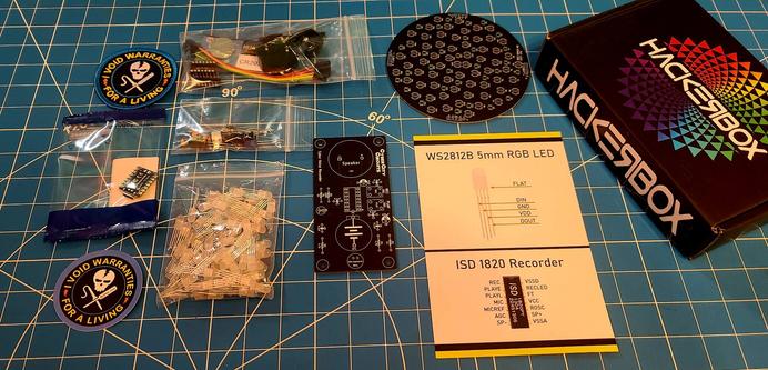 Contents of the May 2023 Hackerboxes subscription. It's entitled Fibonacci and appears to be an LED project that does Fibonacci patterns. An RP2040 is included along with a patch and sticker declaring that "I Void warranties for a living" with a skull logo. A second PCB has a speaker, battery, and switch labels for record and play.
