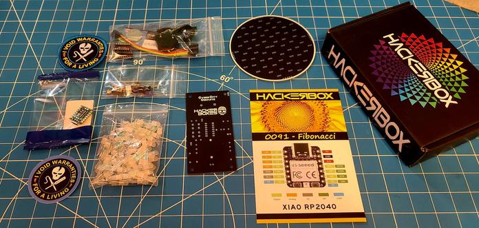 Contents of the May 2023 Hackerboxes subscription. It's entitled Fibonacci and appears to be an LED project that does Fibonacci patterns. An RP2040 is included along with a patch and sticker declaring that "I Void warranties for a living" with a skull logo. A second PCB has a speaker, battery, and switch labels for record and play.