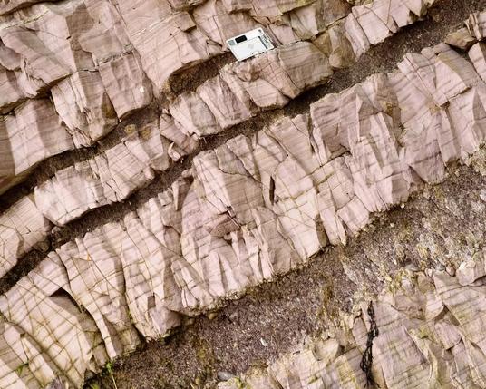 This photograph (scene is about 60 cm tall) shows interbedded argillite (smoother, pinkish, laminated rock) and tuffaceous (volcanic ash) layers that have been altered and weathered back from the rock face. The scale at top is about 5 cm tall. My photo.
