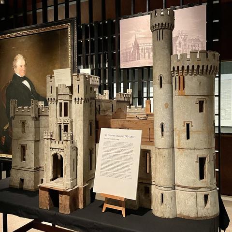 Large model of a neo-gothic big house in the style of a castle with towers and crenellations. An informative panel rests on a mini-easel in front of the model. The rest of an exhibition is partially visible behind the model.