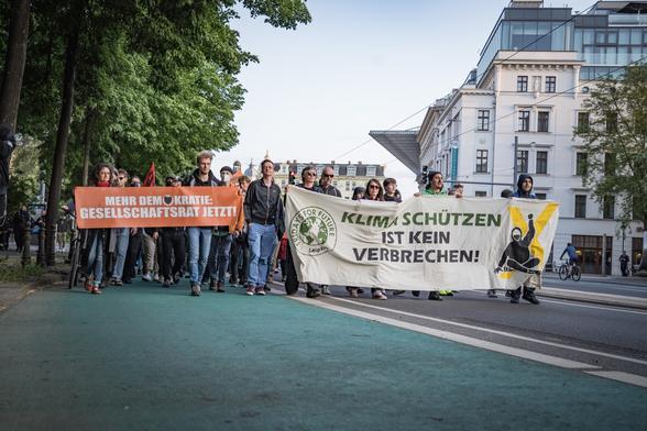 Auf dem Frontbanner der Demonstration steht "Klimaschützen ist kein Verbrechen!". Daneben laufen Aktivist*innen der Letzten Generation mit einem Banner "Mehr Demokratie: Gesellschaftsrat jetzt"