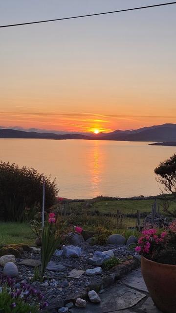 The sunrise of the bay in portrait mode. Roses and geraniums on a terrace, then the water of the bay with an orange sunbeam from the just rising orange sun at the endvof the bay