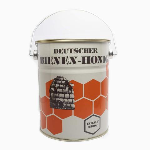 Tin bucket with lid and wire handle, with a pattern of hexagons, a vintage illustration of a basket-style bee hive, labelled "Deutscher Bienen-Honig" ("German bee-honey"; for some reaons "Bienenhonig" is a normal label for honey, though I don't remember coming across any other animal replacing "bee"...)