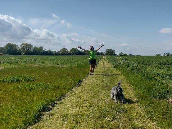 Lady standing on a freshly cut grassed bridleway through open fields with small grey dog in foreground