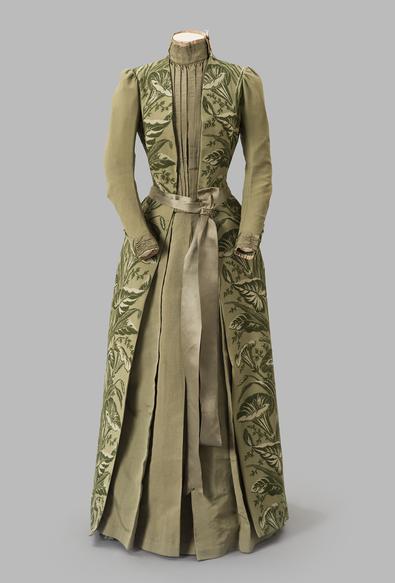 A sage green hostess's gown with a wide satin sash. The side panels from bodice to floor are embroidered in darker green in the shape of calla lilies and leaves. It has a high neck and pleats at the front of the bodice, as well as long sleeves.