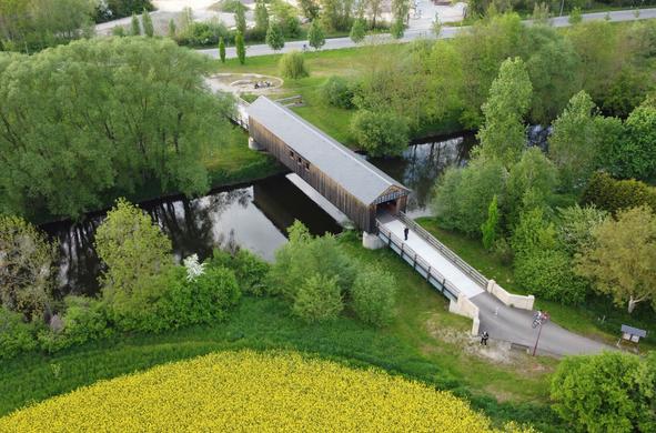 Aerial view of a small covered bridge crossing a narrow river. Both banks are framed by plenty of lush greenery: grassland and many trees. A glaring yellow rape field begins at the lower edge of the picture. In the middle of the bridge entrance, a photographer stands taking pictures through the bridge. On the opposite bank behind it are a few benches where several tourists are taking a rest. A cyclist is riding on the road behind. The scenery seems remarkably peaceful.