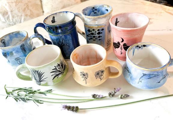 Seven mugs on a marble table. The mugs are painted in pastel colors and have different black designs on them. the designs are agave, fish, jellyfish, daisy flowers, dolphins, black birds and trees.