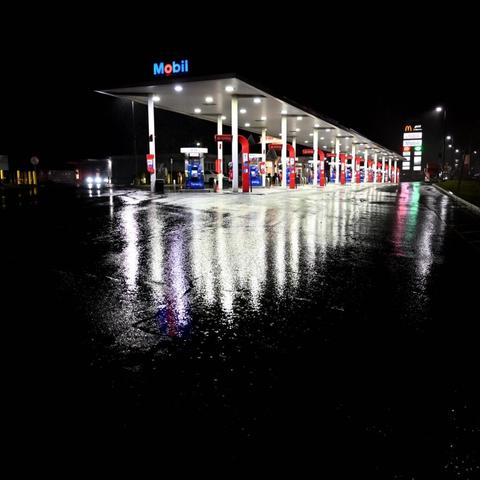 On a dark night, rain-soaked asphalt reflects a brightly lit highway gas station.