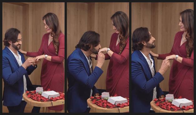 A montage of three adjacent shots shows Sourabh Raaj Jain and Ridhima ma'am together in a romantic moment. Sourabh is to the left of the frame, wearing a blue serge suit and a white shirt. Ridhima ma'am stands to the right of the frame, wearing a crimson wrap dress and a delicate silver watch. Sourabh is down on one knee in front of Ridhima ma'am, her hand in his. In the left-hand image he is placing a diamond ring on the fourth finger of her left hand. In the centre image he is kissing her hand while looking up at her. In the right-hand image he is looking up at her.