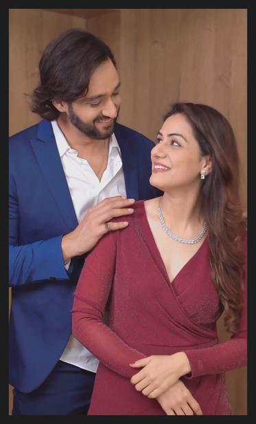 Ridhima ma'am smiles back at Sourabh Raaj Jain, standing behind her, as he places his hands gently on her shoulders. She is wearing a crimson wrap dress, diamond earrings and a diamond necklace. He is wearing a blue serge suit and a white shirt, and is smiling down at her.