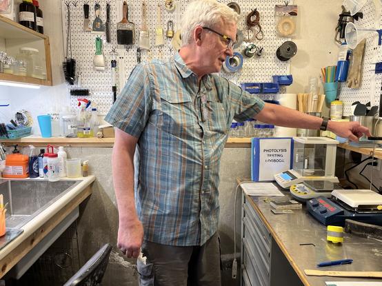 Image shows a man with white hair wearing a greenish blue shirt with red stripes standing in a small room with a sink behind him and in front of him are all sorts of weights on a desk to measure chemicals. Behind him are paint brushes and asorted tools and not shown are a bunch of chemicals