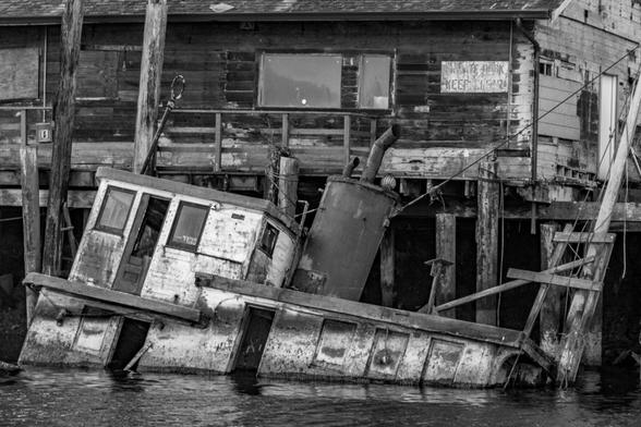 Sunken Boat In Noyo Harbor In Black And White

This is an image of a boat that sunk during a strong winter storm on California's North Coast. 
This was captured during high tide so most of the hull is under water leaving mostly just the cabin exposed. 
The cabin is missing a couple of doors and the finish is weathered with paint peeling off of the wood. 
An old warehouse sits behind the boat on the docks. The building is weathered as well and reflects the decay seen on the boat.