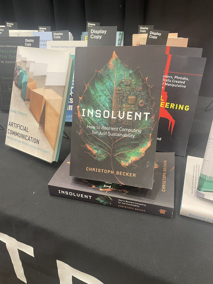 A book stand at a conference exhibition table, showing at the center a book titled INSOLVENT: How to Reorient Computing for Just Sustainability by Christoph Becker. The cover features a green leaf enmeshed with digital circuitry, rotting at the edges in fiery copper on a black background.