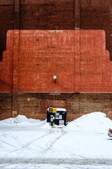 A brick wall with a very wash out advertising painted on, and a shape of what look like a missing building. It its foot a parking meter and the ground is covered in snow. To the right the tail lights of a car make it to the frame.