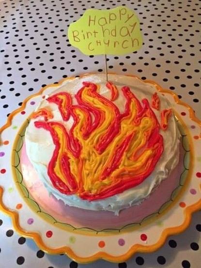 Iced cake, white nase, red and orange flame in centre. Flag with words "Happy Birthday Church" written by a child