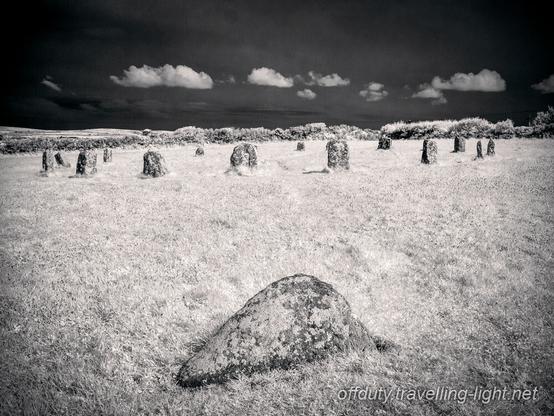 View of a stone circle with a single stone in the foreground, Beyond the circle is a hedge and clear black sky with a line of small white clouds.