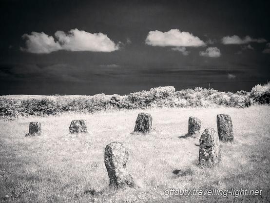 View of a part of a stone circle. Beyond the circle is a hedge and clear black sky with a line of small white clouds.
