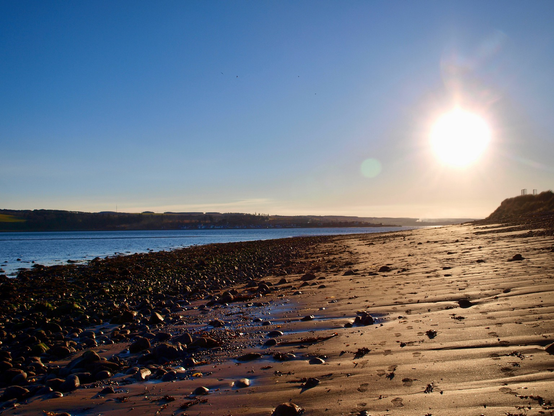 Blue sky with a starburst sun, silhouette of a town across the icy blue water, pebbles at the shore line and ridged sand towards the right edge of the photo