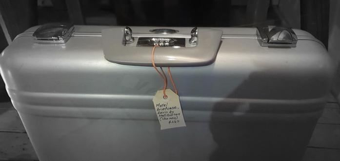 A briefcase for sale in a shop window. A handwritten tag reads: "Metal briefcase. Zero by Halliburton. $250"

The briefcase has a 3-digit combo lock. Once unlocked, you flip two clasps on either side. Only one side has a handle.
