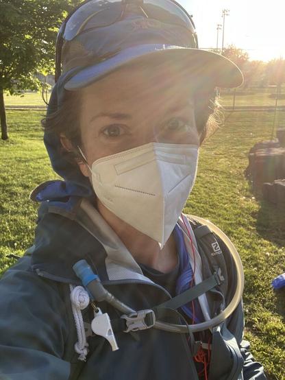A selfie before a race of a middle aged white woman in a blue hat with sunglasses on her head, a kn95 mask, a blue shell, blue hoodie, and a hydration tube and whistle on the straps of a green backpack. She is backlit by the morning sun and smiling