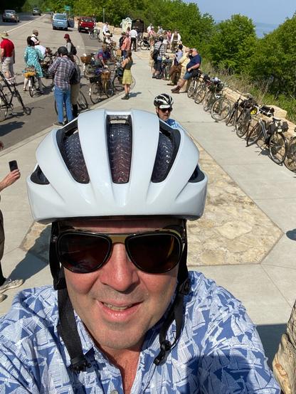 An old man taking a selfie while wearing a bicycle helmet. Throngs of English three speed bicycles are parked at a scenic lookout behind him, while their owners mill about