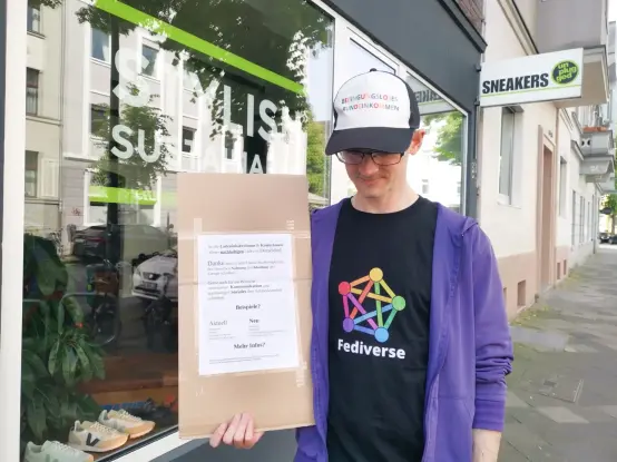 Mensch (Mann) steht mit einem Pappkarton welcher eine Botschaft für nachhaltigeres Handeln aufzeigt, auch in den Bereichen Kommunikation und Soziales. z.B.: aktuell Instagram, neu Pixelfed. aktuell Twitter, neu Mastodon. Aktuell Bürgergeld /Hartz4, neu Bedingungsloses Grundeinkommen. Aktuell Extrinsisische Motivation, neu Intrinsische Motivation. Vor einem Sneaker Store in Düsseldorf Flingern Names Sneakers Unplugged. Am Schaufenster steht "Stylish Sustainable"