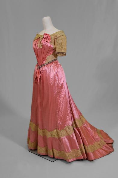 pleated pink satin silk gown with lace sleeves, V waistline, and floral embellishments. Slight train and wide, almost boat-neck cut at top.