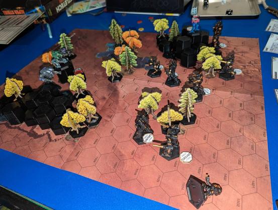 Several Battlemechs on a hex grid surrounded by 3d printed hills and cardboard trees creating a fleshed out battlefield.