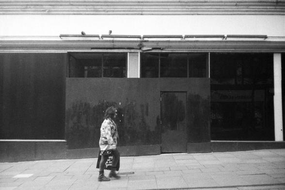 A 35mm black and white film photograph of a shopper passing an empty shop front in Wigan.