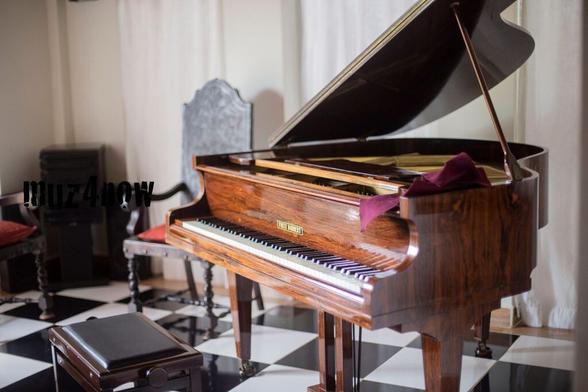 A grand piano with a natural wood finish sits in a room with a black and white checkerboard floor. The lid is up on the piano and there is a piece of purple felt on top of the right edge of the instrument. Besides the empty piano bench, there are at least two other wooden chairs in the room.