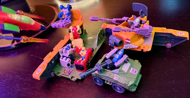 The Anti-Aircraft Base deployed in base mode on a desk, filled with Micromasters. The race car patrol is present sitting down in the main part of the base, except for Tailspin who is manning a missile emplacement. The Decepticon Micromasters are manning some guns, aiming for Thundercracker in garish Action Master/Shattered Glass colors who in jet mode who is sweeping in from the edge of the image