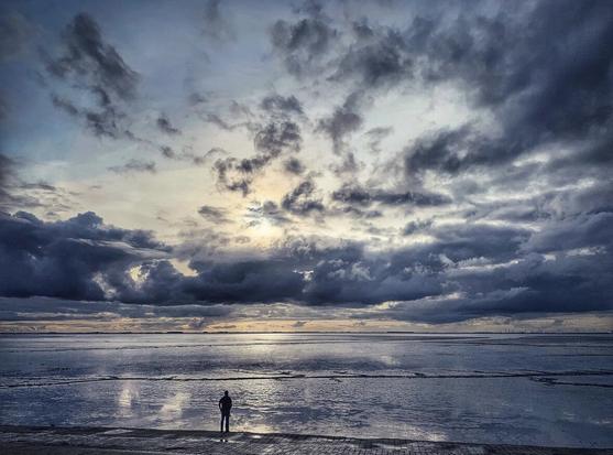 A person in the distance looks out into the vastness of the North Sea coast, accompanied by a dramatic sky with clouds and sunset.