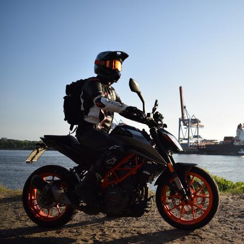 A motorbike rider on his KTM 390 Duke. In the background there is a channel of water. On the other side there are loading cranes and containers.