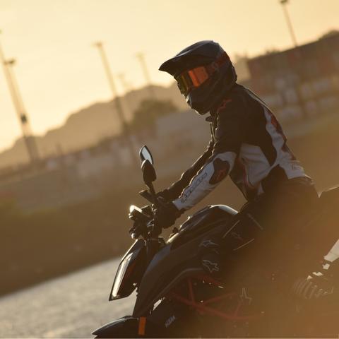 A motorbike rider on his KTM 390 Duke. The whole picture is golden due to the sun setting. The background is blurry, but a channel of water, containers and a crane can be made out.