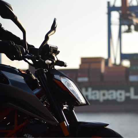 The front of a KTM 390 Duke. In the background there is a channel of water. On the other side there are loading cranes and containers.