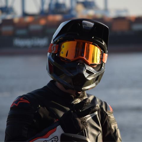 A motorbike rider showing the peace sign. In the background there is a channel of water. On the other side there are loading cranes and containers.
