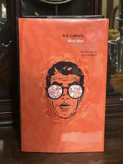 A bright orange cover with a line drawing of a man's head facing toward the viewer.  He's wearing an oversized pair of glasses with circular lenses.  The lenses are swirling with white, pink and blue, and you cannot see his eyes.