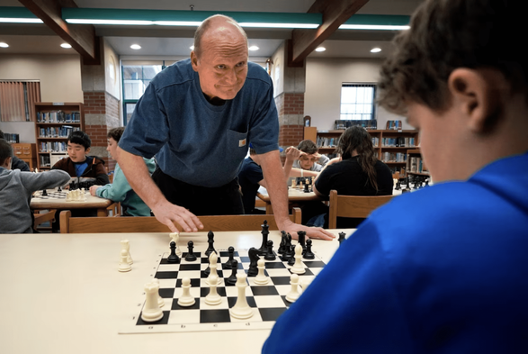 An older man (facing the camera) plays chess with a 6th-grade boy (over the shoulder)