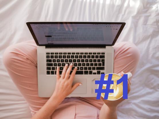 Kobieta siedząca po turecku na łóżku i trzymająca laptopa na kolanach. Lewa dłoń położona jest na klawiaturze, a prawa trzyma kawę ze wzorem. Kadr jest z góry (nie widać twarzy kobiety), a na kawie jest napis #1