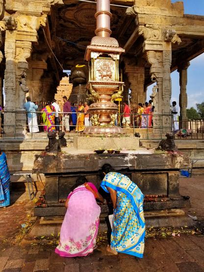 Women in colorful saris placing clay bowls of water at the base of a gold and stone pillar.