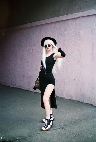 White hair girl in a black dress wearing black hat and rounded sunglasses. She poses so as her left leg almost fully exposed and her left hand slightly touches her hair.