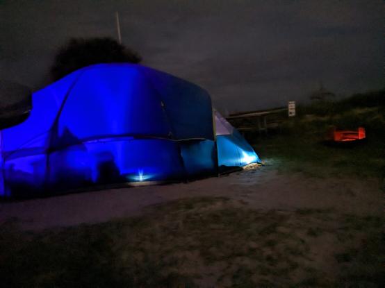 A tent for camping is illuminated from the inside at night with a fire pit glowing to the right.