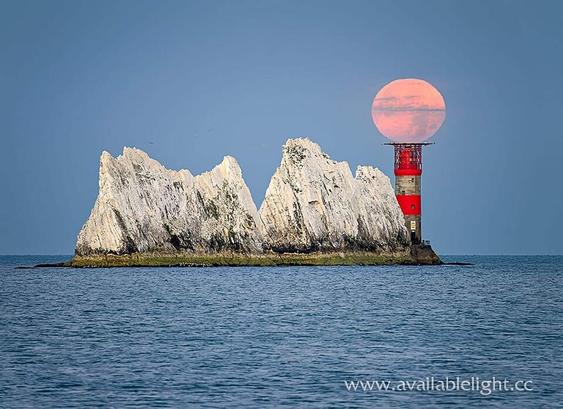 Two huge chalk stacks with a red and white striped lighthouse at the end. The setting full moon appears to be balancing on top of the lighthouse. The colour of the sea and sky is a muted blue, almost a denim like colour owing the sun being on the verge of rising.