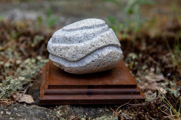 A carved Granite oval shaped stone 3.5 inches long by 3 wide by 2 high (9cm by 7.6 by 5) seen by side view. The top has been carved in two layers resembling a mountain plateau, the top layer thinner than the second. A deep groove in a wavey pattern has been carved along the bottom 1/3 of the stone.