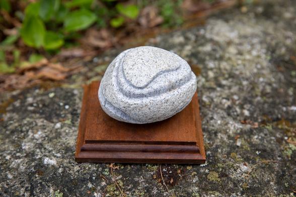 A carved Granite oval shaped stone 3.5 inches long by 3 wide by 2 high (9cm by 7.6 by 5) seen looking down. The top has been carved in two layers resembling a mountain plateau, the top layer thinner than the second. The top layer is kidney shaped while the second layer is more oval. A deep groove in a wavey pattern has been carved along the bottom 1/3 of the stone.