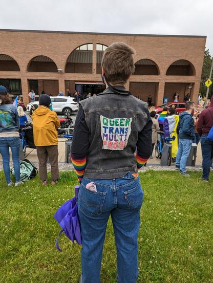 Person wearing a denim vest with a Queer Trans Multi Proud patch in various colored lettering