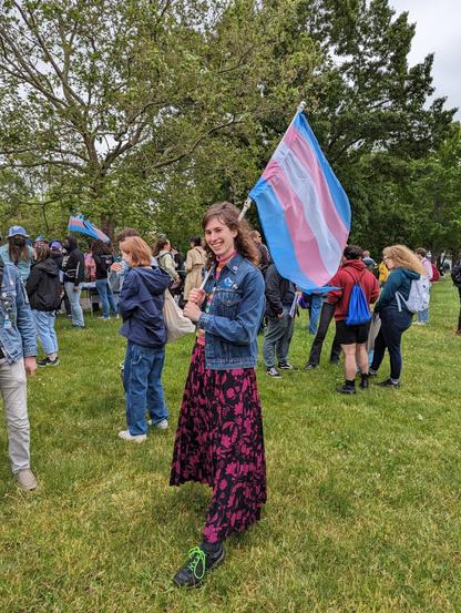 Person in a fuscia and black colored skirt and jean jacket smiling and holding a trans flag (blue, pink, white stripes)